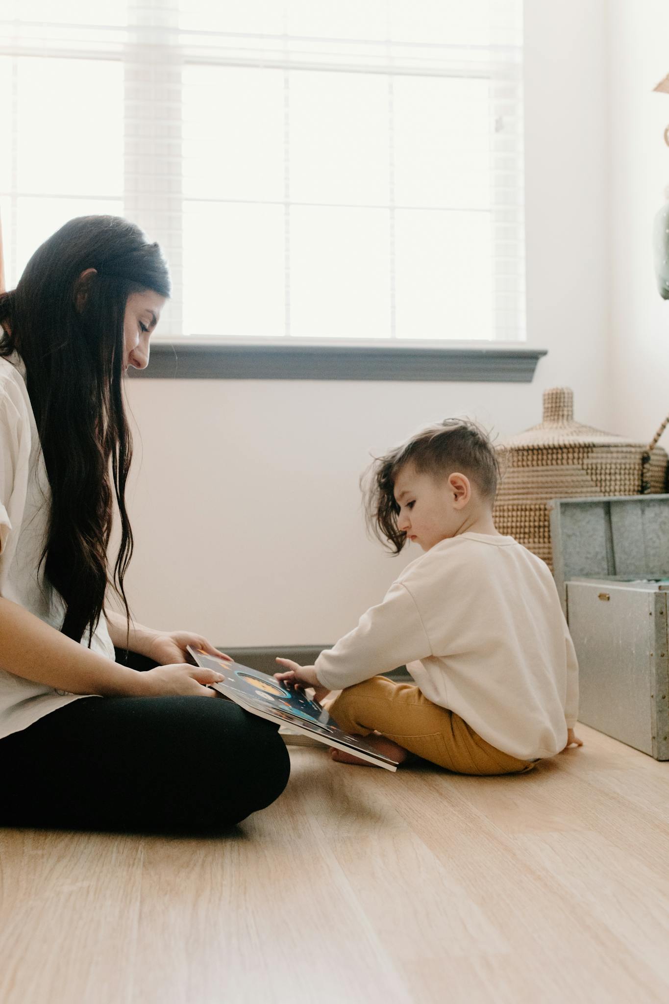 A mother and child sit on the floor, reading a book in a cozy indoor setting.