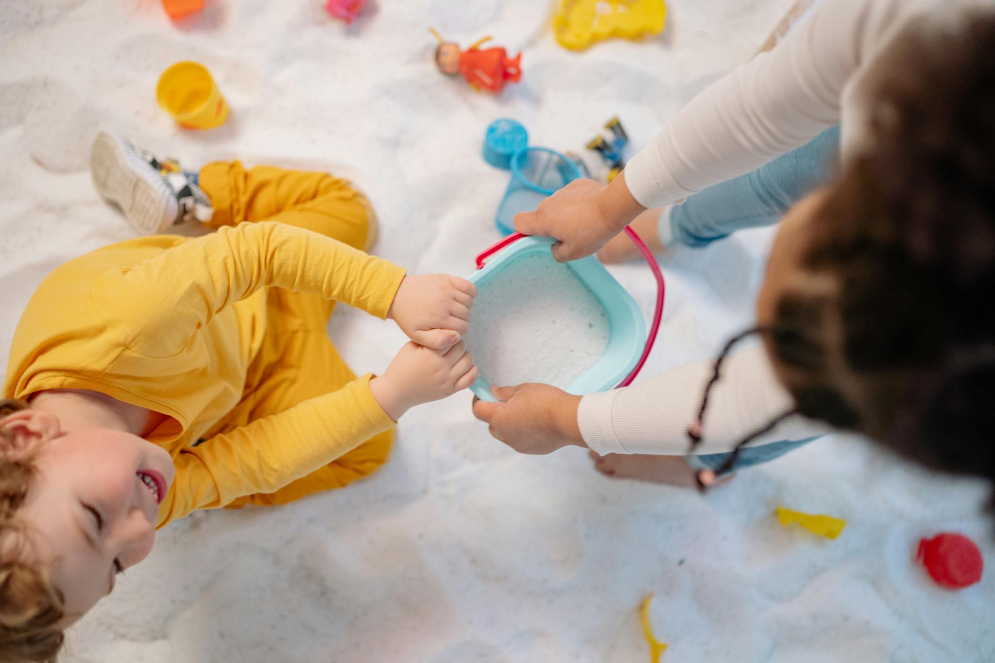 Two children play with toys and a bucket on a sandy surface indoors.